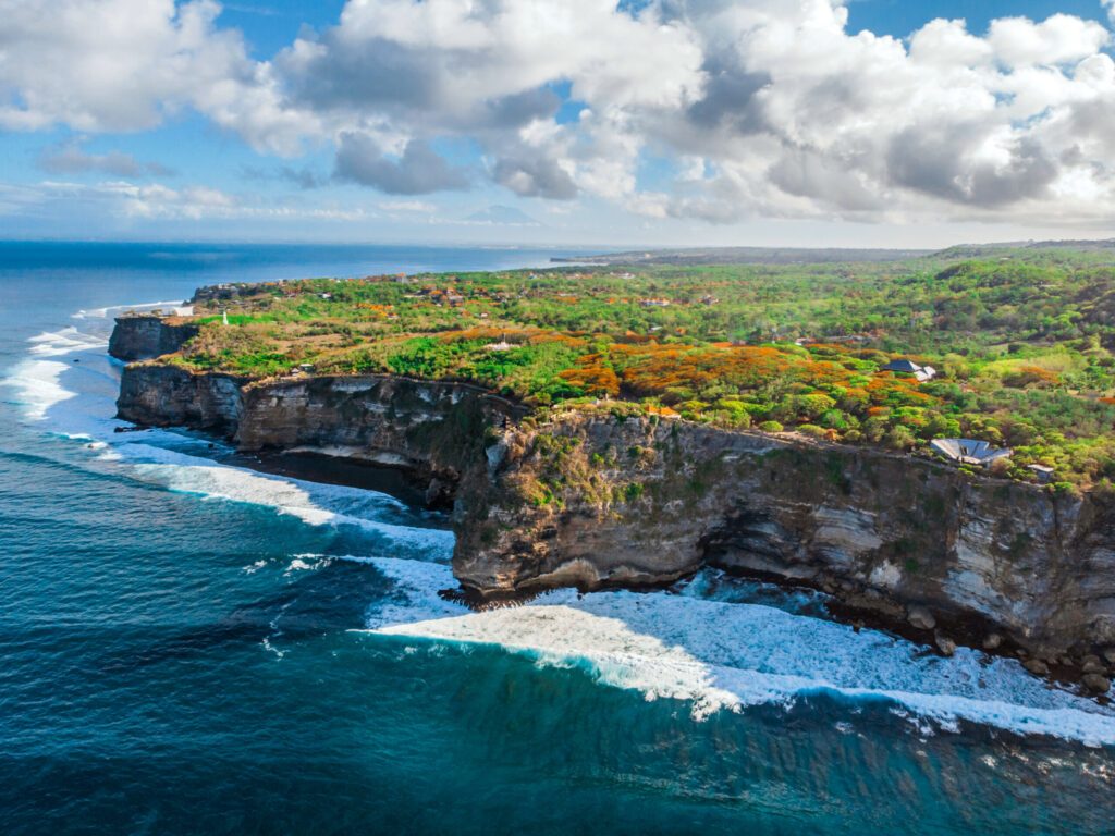 The sheer cliffs of the southern coast of Bali are washed by a clear azure ocean. Bukit Peninsula aerial view. Uluwatu temple on the rock.
